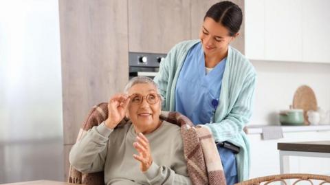 An older woman with very short grey hair and round glasses. She is sitting at a table and has a blanket over her shoulders. She is looking up behind her to a carer. The carer has dark hair tied back and she is wearing blue scrubs and a mint green cardigan. They are in a kitchen