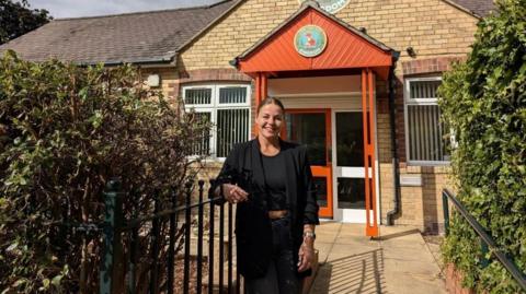 Emma Fullwood is standing by some railings in front of a nursery which has a sign My Little Kingdom above the door. It's a modern one-storey building with a gable end