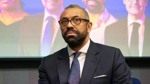 James Cleverly looking serious while sat on stage. He is wearing a black suit jacket with a white shirt and blue and red spotted tie.