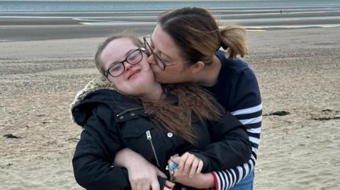 Emily McAllister, wearing a navy jumper, hugging and kissing her daughter Keira, who has down syndrome and is wearing a black coat, as they stand on a sandy beach. 