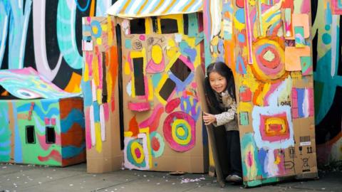 A little girl with long black pony tails opens the door of a cardboard house painted with colourful shapes.