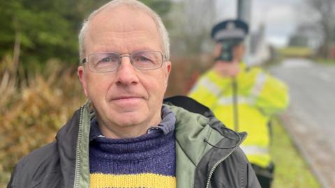 A man with grey hair and glasses in a blue and yellow jumper and green jackets stands in front of a polic officer with a speed camera in his hand