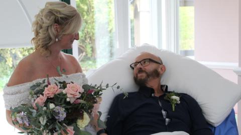 A blonde woman in an off-the-shoulder wedding dress holding a bunch of pink and cream roses and purple flowers, with her face angled towards a man in a black shirt propped on a pillow on a bed. He is looking to his right back at her. He has a cream flower in his lapel. He is wearing glasses and has a dark beard. The pair are in a room with pink walls and glazing behind them.