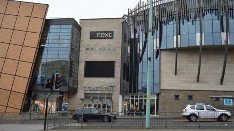 A general view of Drake Circus shopping mall in Plymouth. Cars are travelling along the road in front of the mall. It has large glass windows. Shop logos including Next and Primark are on display.