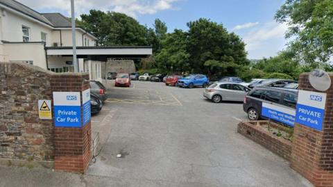 The Chadwell Centre in Paignton. The car park outside the white and cream-coloured building is filled with vehicles. A brick wall is on the perimeter of the car park. There are blue and white NHS signs saying the car park is private. Trees are dotted around the opposite side of the car park.