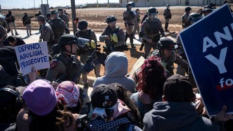 a group of protesters, holding anti-ICE signs, stand in front of a group of state troopers on a desert road