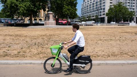 A man wearing a white shirt and black trousers rides a lime bike in the centre of the picture through a park in London with yellowed very dry grass. Traffic and buildings can be seen in the background