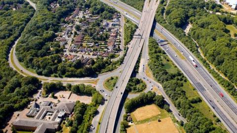 An aerial image of a road surrounded by cars and trees. The road has several cars on it.