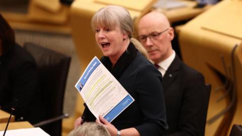 Shona Robison holds a piece of paper up in the Holyrood chamber. Her blonde hair is back in a ponytail and she wears a black blazer. John Swinney can be seen behind her - he wears a white shirt, dark red tie and a black suit jacket.