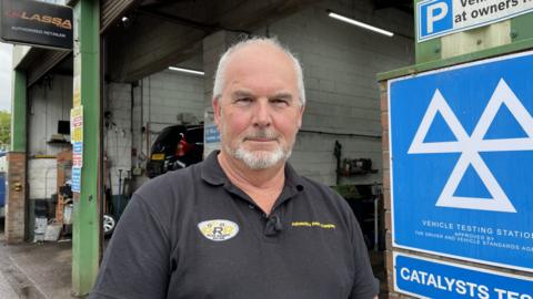 A man with white hair and a beard stands outside a garage, looking into the camera. He is wearing a black T-shirt with yellow stitching that reads 'Automotive Repair Company' below his left shoulder, and below his right shoulder is a yellow and black logo stitched onto a white oval background, with three Rs together and the words 'Road Runner Racing' below.