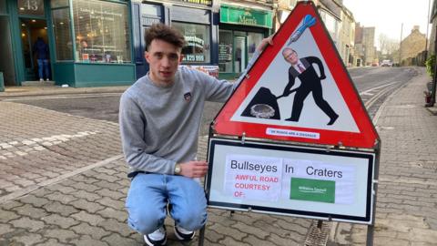 Ben kneeling down next to a spoof roadworks sign, outside on a street.