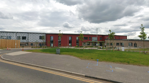 A two-storey building is set back from the road, beyond a grassy area with newly planted trees. The left of Maybury Primary school has a white and grey fascia, with a dark red fascia for the rest of the building and a single storey brick-built wing to the right.