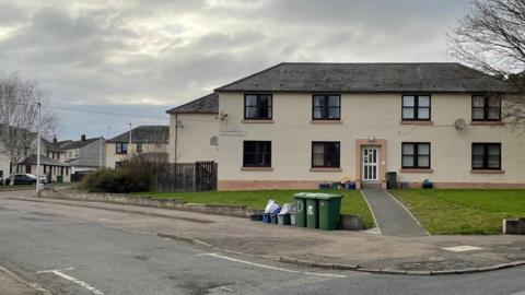 A residential street, with a focus on one house on the corner. It is large with white walls and a grey roof. The front garden has trimmed grass and a low grey wall with bins sitting by the wall