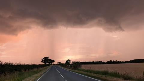 A single carriageway country road with fields on either side. There is a large, dark storm cloud covering the top of the image and there is a pinkish light being cast by the sun which is hidden. There are trees in the distance which look black due to the shadow.