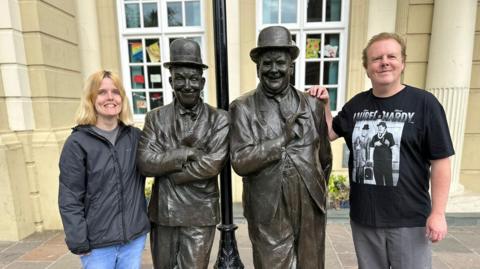 Rebecca Sills - a young woman with blonde hair, wearing a waterproof coat, and Dean Sills - an older man wearing a black Laurel and Hardy T-shirt. They are standing next to statues of Laurel and Hardy. 
