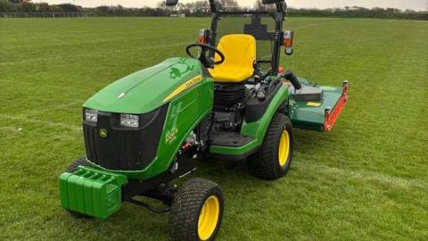 A picture of a green husqvarna ride-on mower. The mower is on what looks like a football pitch and there are trees and bushes in the background.