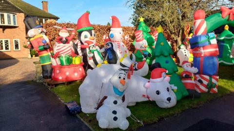 A large collection of about 20 brightly coloured inflatables on the front verge outside a detached brick house. There are a variety of snowmen, Christmas trees, presents and other decorations.