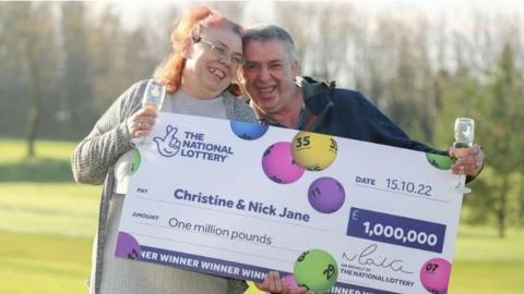 The couple smiling and holding their oversized cheque which has their names and the amount printed on it. Christine has long tied up pink hair she is wearing a grey dress and a grey cardigan. Nick has short grey hair and is wearing a blue jacket. Both are holding champagne flutes which are partially filled.  