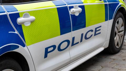 The side of a white police car with blue and yellow panels along the door and the word 'POLICE' written in blue.
