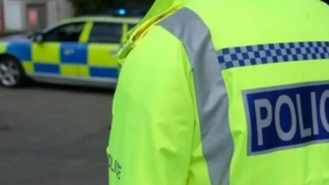 The back of a yellow hi-vis police officer's jacket on the right. It has the words 'POLICE' on the back. On the left is a blue and yellow police car.