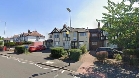 A Google Street View image of Coalway Road Surgery, a cream and black Tudor-style building on a residential street in Wolverhampton. A small car park with two vehicles sits in front of the surgery, bordered by low hedges and shrubs. Semi-detached houses line the road to the left of the practice.