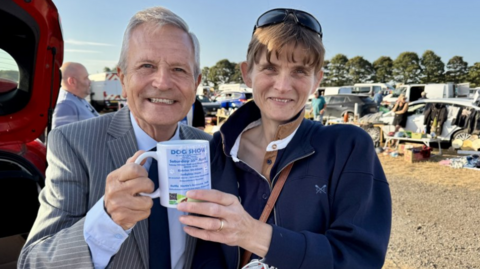 Peter Levy standing next to a woman in a blue jacket. They are holding a mug advertising a dog show. Both are smiling. There are market stalls in the background.