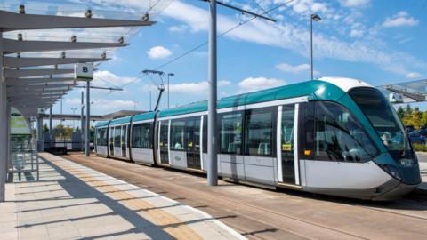 A silver and green tram at a stop on a sunny day.
