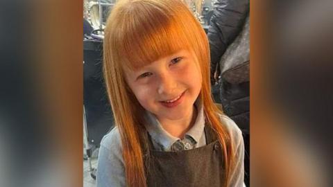 Lila with ginger hair and grey pinafore and light grey top smiling.