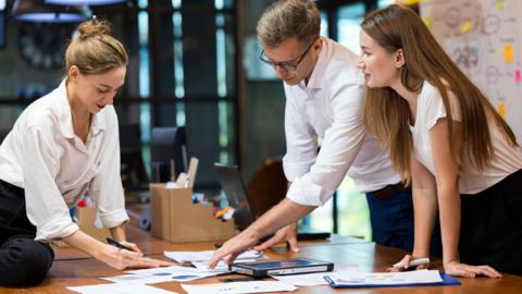 Three people lean over a desk looking at a paper with data graphs on it, holding pens and in discussion.