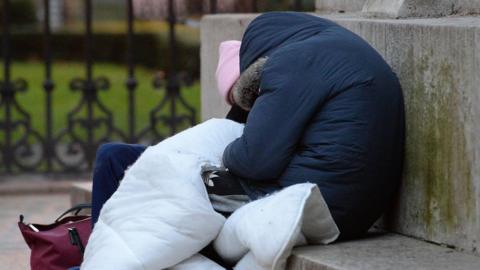 A person appearing to be homeless, wrapped in a hooded blue coat and holding a duvet. They are sat on the steps of a plinth in a square.