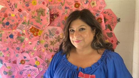 A smiling woman stands in front of a pink quilt made up of hexagonal patches with flowers on them