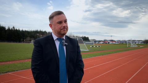 Colin George Moireasdan na sheasamh aig oir pàirce agus là math air a chùlaibh.