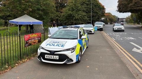 Two police cars are parked on the pavement outside Cripplegate Park and a blue tent has been put up in the park itself. 