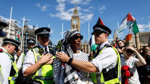 Police officers hold a woman protester back. People hold Palestinian flags around them and Big Ben's clocktower is visible in the background.