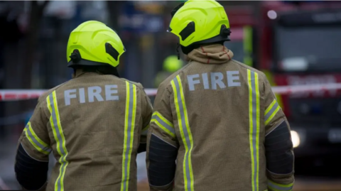 Two firefighters wearing yellow helemet and a fire jacket. They have their backs turned away revealing the word "fire" in bold white lettering.