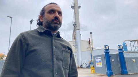 A man in a dark green jacket standing outside the blue stone gateposts of the rendering factory
