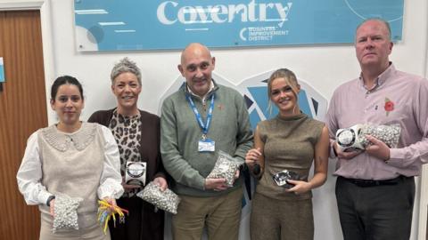 Three women and two men standing in a line, holding a variety of small items, including clear plastic bags containing small white objects