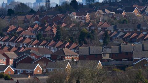 A wide view across rows of red-brick residential rooftops in Dudley, with the Birmingham city skyline visible in the hazy distance. The BT Tower and construction cranes rise above the horizon behind a mix of suburban housing, trees and tower blocks. 
