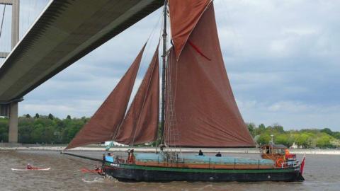 Spider T sailing under the Humber Bridge, It has red-coloured sails and rigging. There is a person in a canoe nearby.