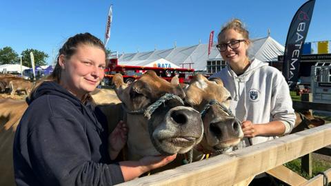 Two women in hoodies stand either side of two brown cows behind a fence at the Driffield Show