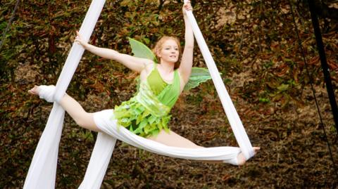 A woman wearing a light green dress, with green fairy wings, is suspended on white bands doing acrobatics against a rustic background. 