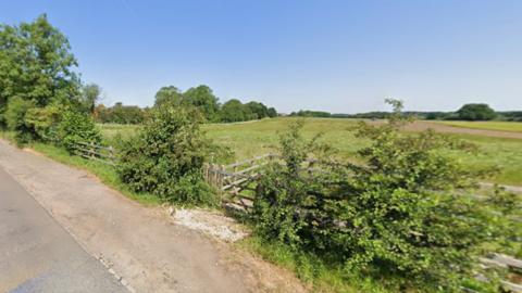 A Google streetview image, showing land north of Fisherwick Road in Whittington. There are fields behind a hedgerow with a wooden gate and fence. There is a road in the foreground, and the scene is set against a blue sky.