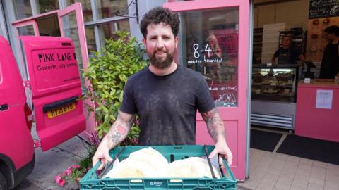 Abe Chirgwin is standing outside Pink Lane Bakery holding a green box full of stotties. The bakery doors and counters are pink. Mr Chirgwin has dark curly hair and a beard and is wearing a grey t-shirt.