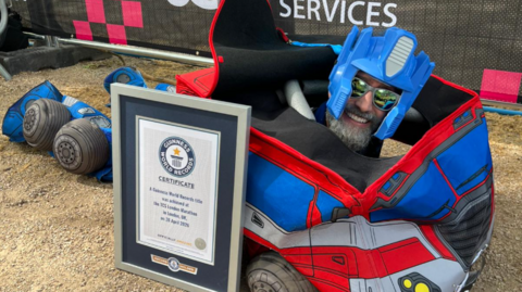 Matt Batchelor poses on the ground inside a large blue and red Optimus Prime costume, with only his head visible. It is covered in a blue plastic helmet, and he is wearing reflective sunglasses. Propped up against his body is a framed Guinness World Records certificate.