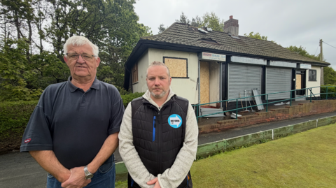 Barrie Bulch is wearing a blue polo shirt and is standing next to Councillor Paul Donaghy, who is wearing a black gilet with a blue Reform UK logo. They are standing on a bowling green in front of the burnt-out club house behind them.
