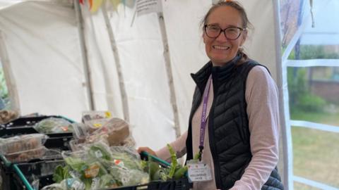 A woman with glasses wearing a black gillet and purple shirt smiling at the camera. She is standing in a marquee in front of crates of food.