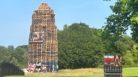 A tall bonfire sits on a grassy green. It has union jacks on it as well as a larger, dark banner. 