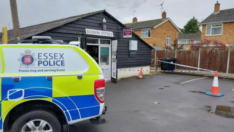 A police car in front of a shop at Baddow Antiques Centre. There is police tape cordoning off the entrance to the building, which is one-storey and black.