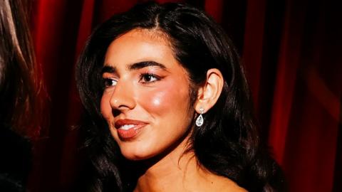 A close-up shot of Izzy Escobar who is stood against a red curtain. She looks to the side and wears sparkling silver earrings and has her long black wavy hear tucked to one side behind her ear