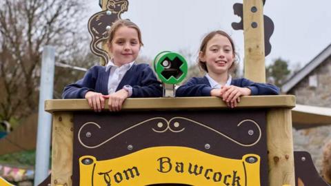 two school girls standing on top of a pirate ship climbing frame.